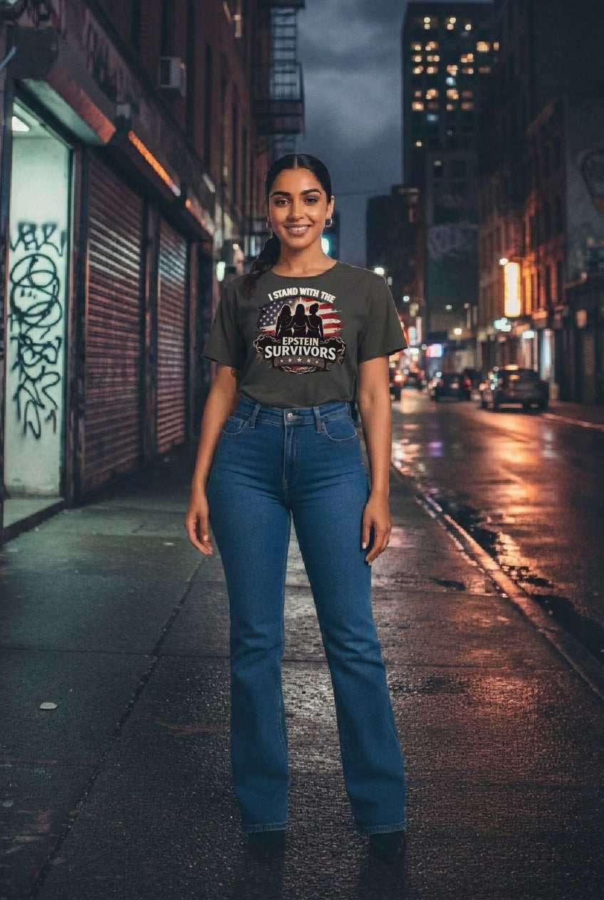 Woman wearing a dark I Stand With The Epstein Survivors graphic tee and blue jeans on a wet city street at night.
